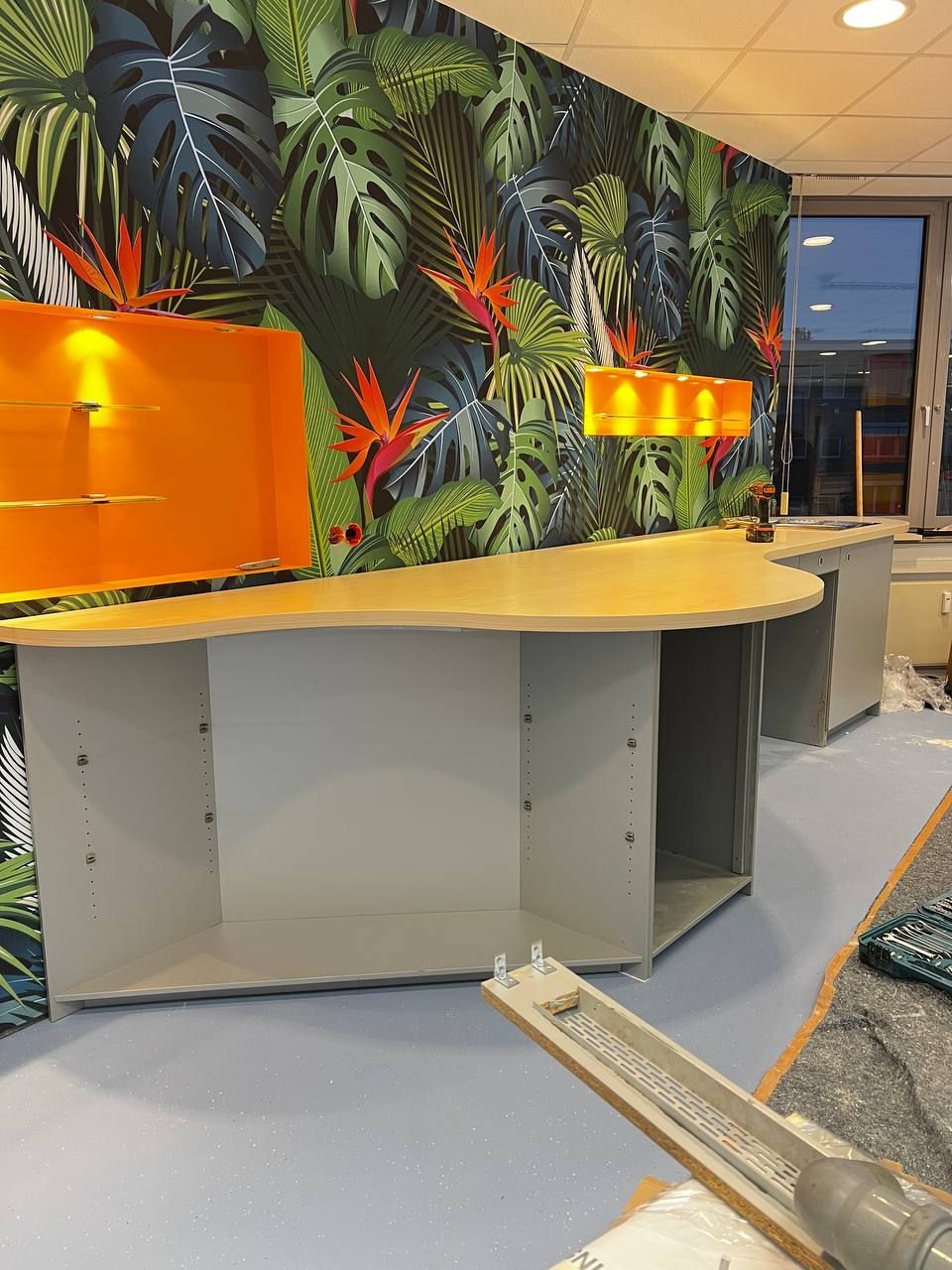 Modernes Büro mit buntem Dschungelmotiv-Tapete, geschwungenem Holztresen und orange beleuchteten Regalen.