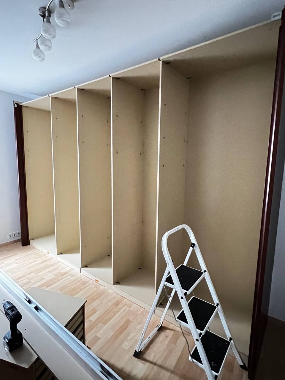 Ein leerer, hölzerner Schrank im Aufbau, daneben eine weiße Leiter in einem Raum mit Holzboden.