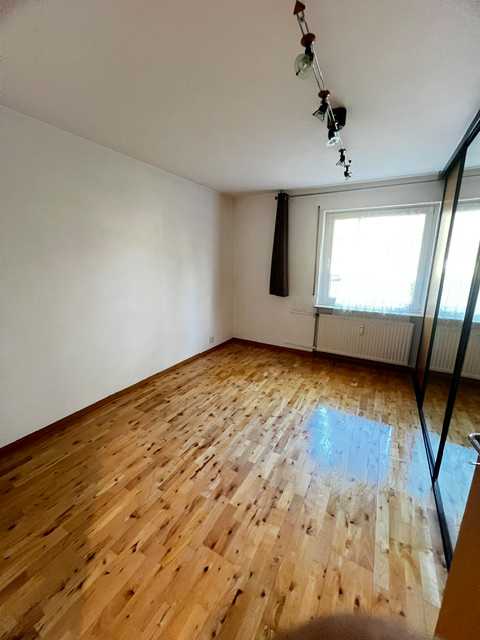 Ein leeres Zimmer mit Parkettboden, weiß gestrichenen Wänden, einem Fenster mit braunem Vorhang und einer Deckenleuchte.